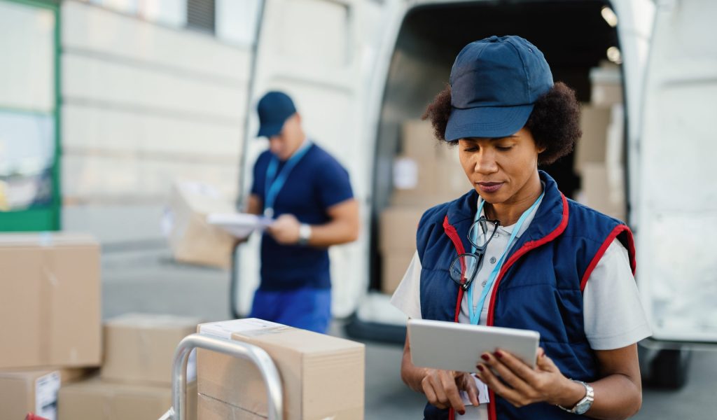 African American female worker making inventory of packages and organizing a delivery schedule on a digital tablet.  Her colleague is working in the background.